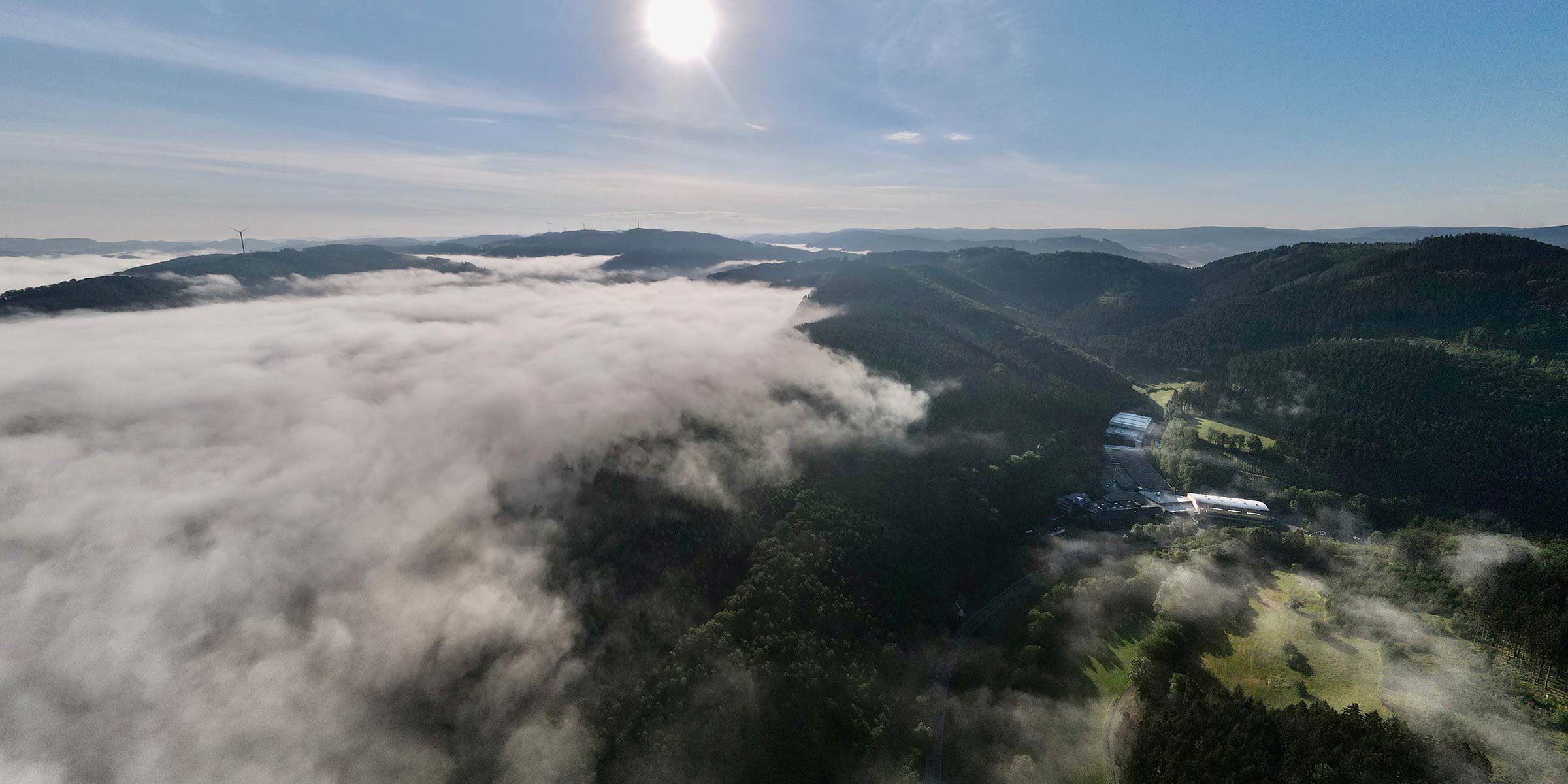 Luftaufnahme des modernen Firmengebäudes im Bonzelerhammer, eingebettet in die idyllische Landschaft des Sauerlands, bei strahlendem Sonnenschein.